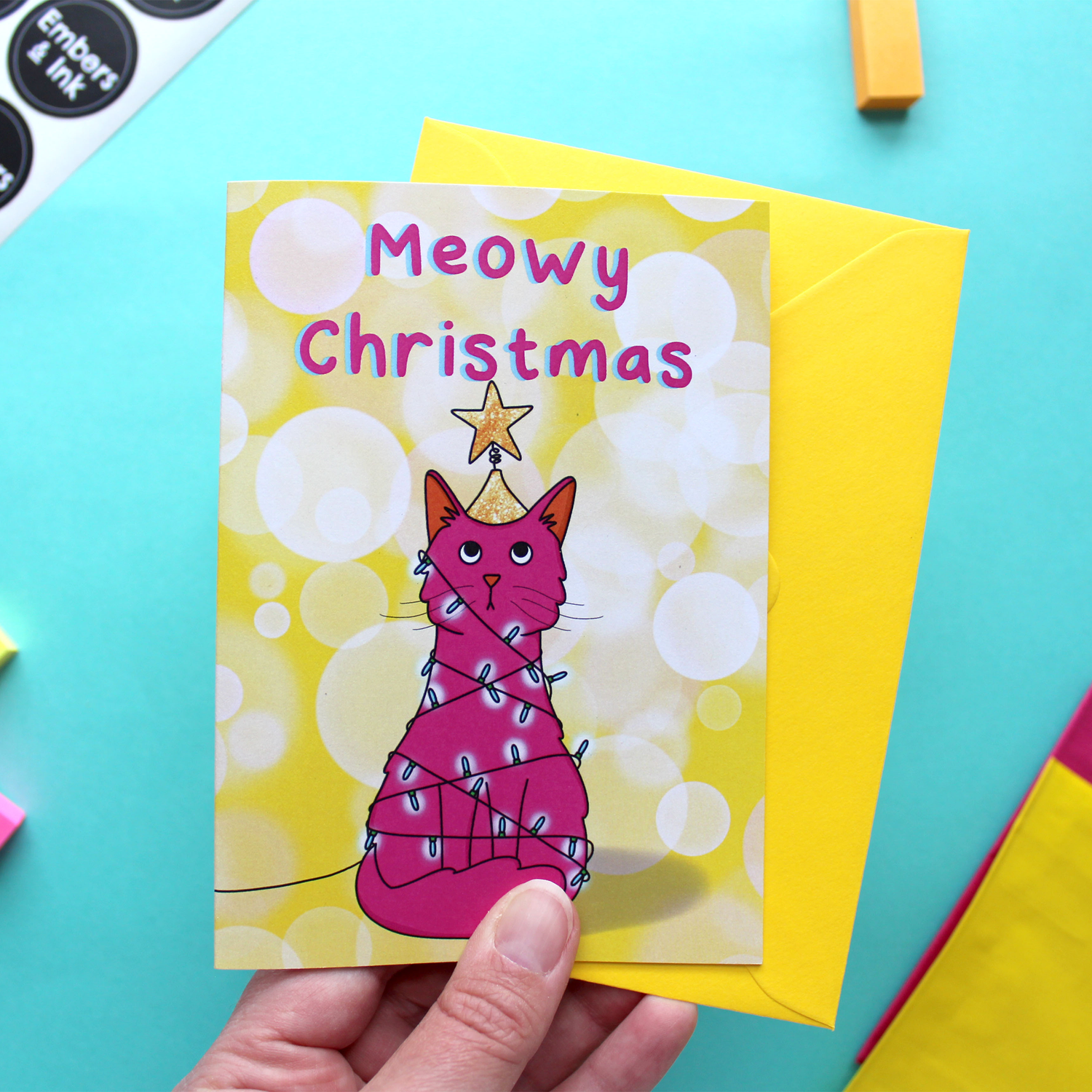 A hand holds a landscape orientated greetings card with an illustration of a black and white car wrapped in lights. Underneath are the words Meowy Christmas