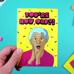 A hand holds up a brightly coloured card featuring an illustration of an oder person looking shocked under the words You're How Old?!'. The text is pink in a fun cartoony font, and the background is yellow with coloured confetti falling down.