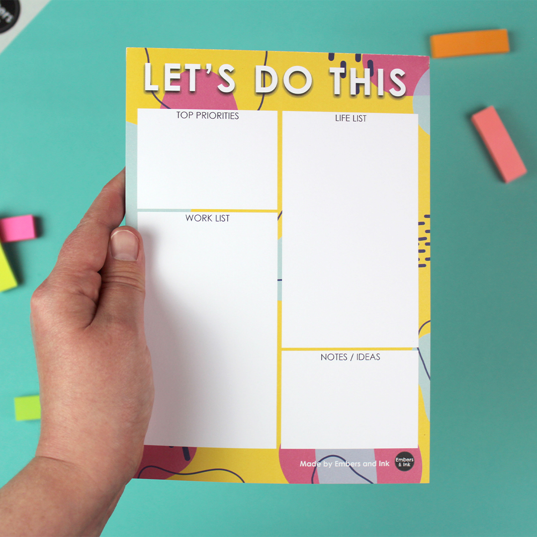 A hand holds an A5 notepad with the words Let's Do This along the top and four white boxes int he middle of a brightly coloured patterned background. The topleft boxes are names Work List and life list (which are bigger boxes of equal size) and 'top priorities' and Notes/Ideas' which are smaller boxes, but the smae size as each other.