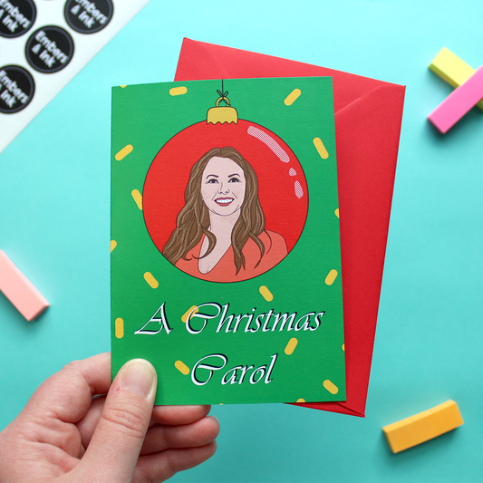 A hand holds a Christmas card that has an illustration of Carol Vorderman (of retro television fame) within a red bauble. Underneath are the words 'A Christmas Carol'. The card has a green background with yellow confetti-type shapes on it.