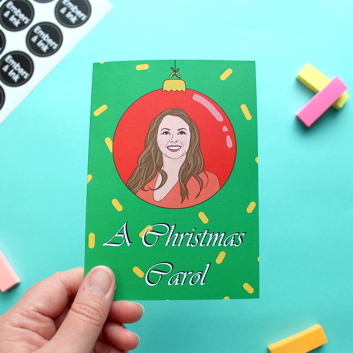 A hand holds a Christmas card that has an illustration of Carol Vorderman (of retro television fame) within a red bauble. Underneath are the words 'A Christmas Carol'. The card has a green background with yellow confetti-type shapes on it.