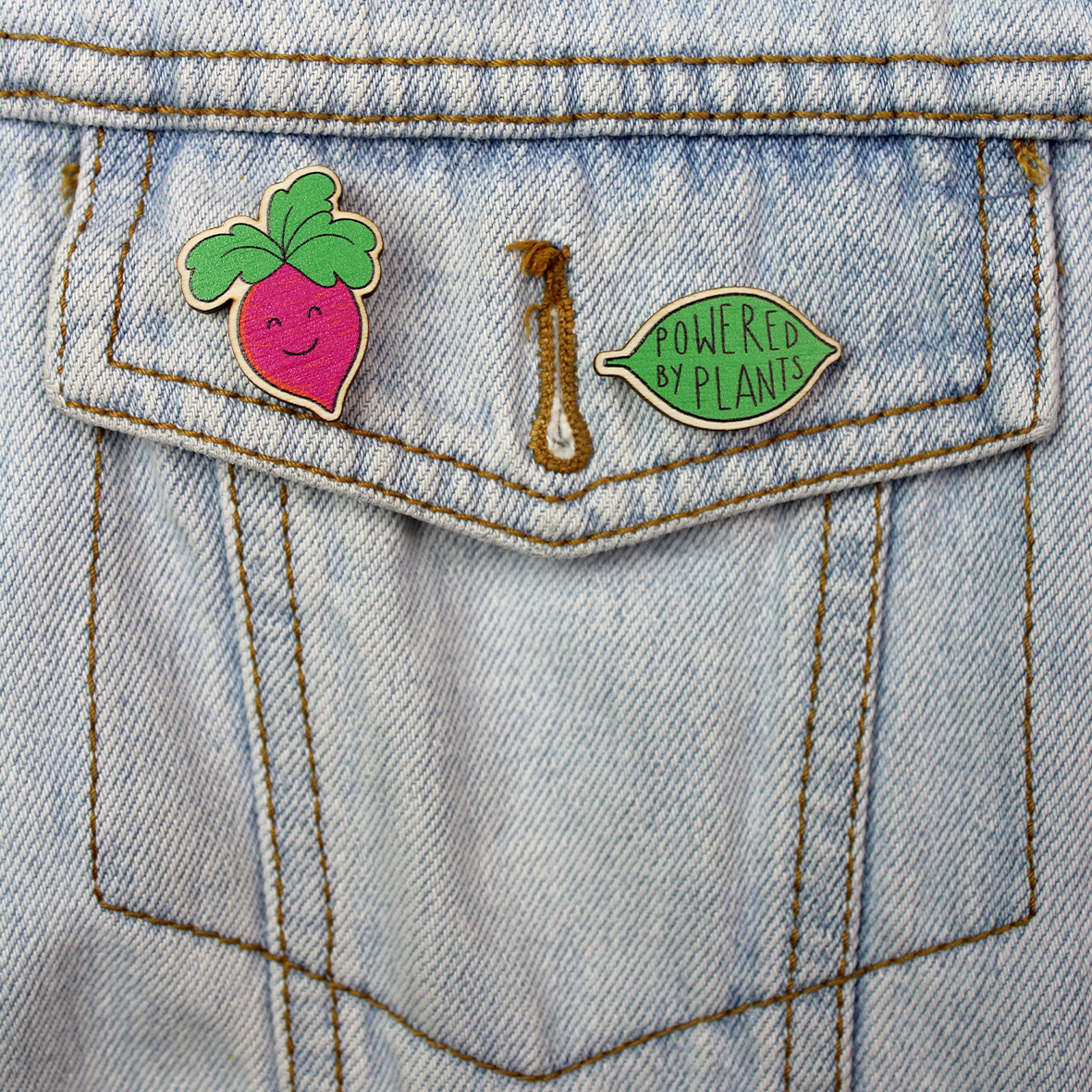 The Powered by Plants wooden pin is shown on a the pocket of a denim jacket alongside another wooden pin badge.