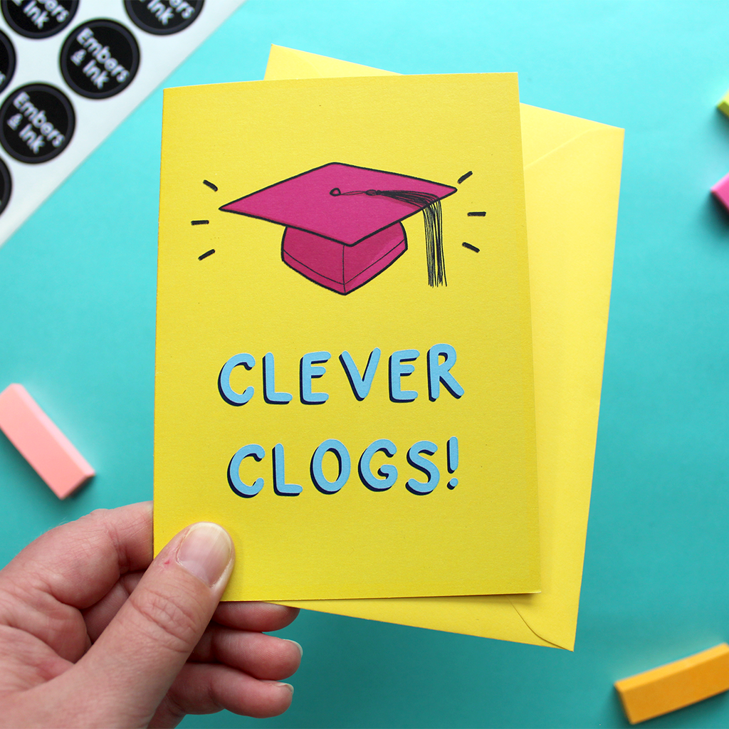 A hand holds a yellow card that features an illustration of a pink mortar board and the words Clever Clogs!