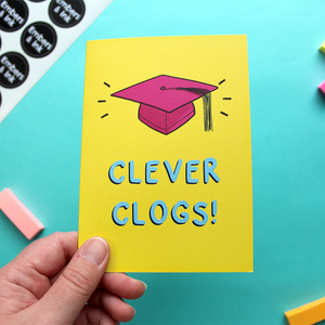 A hand holds a yellow card that features an illustration of a pink mortar board and the words Clever Clogs!