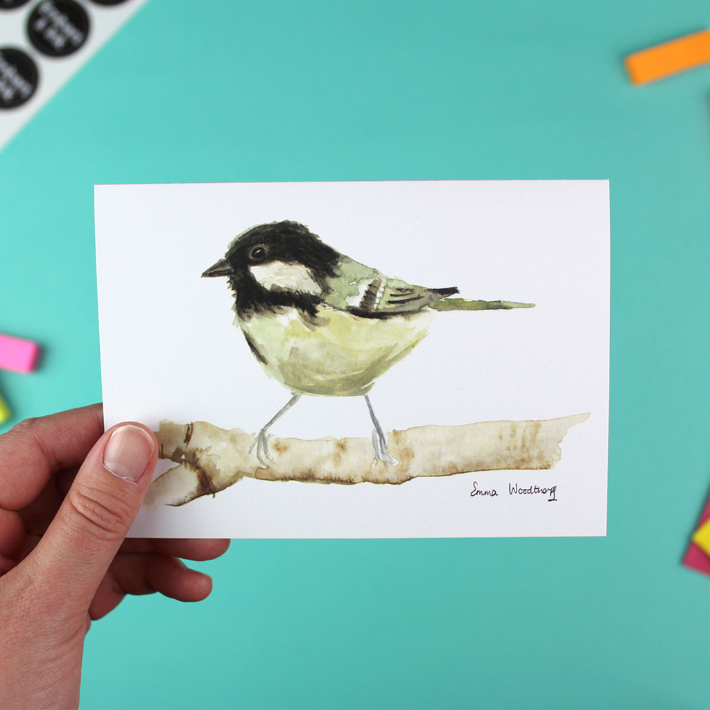 A hand holds up a landscape-orientated A6 greetings card featuring a watercolour image of the birds the Coal Tit.