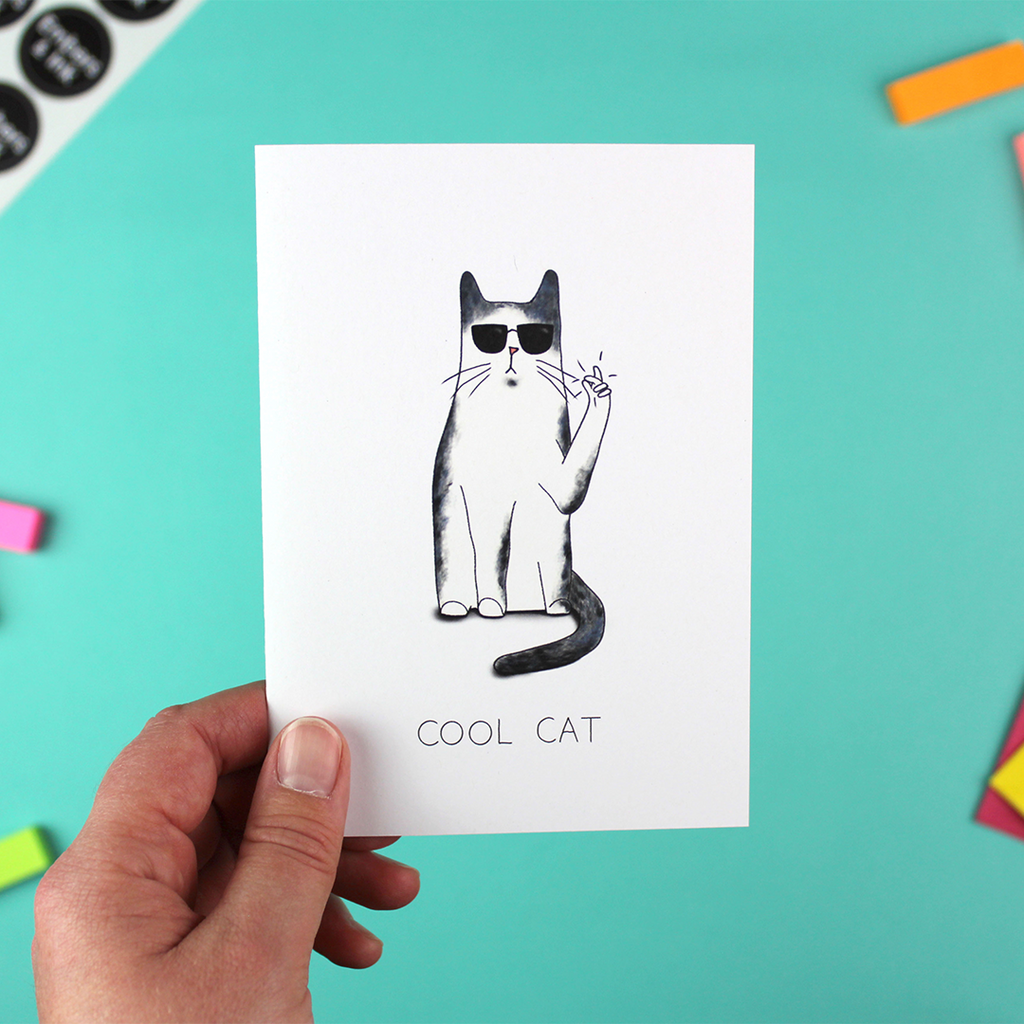 A hand holds up a portrait orientated greetings card with an illustration of a black and white cat that is wearing shades and clicking its fingers. Underneath are the words Cool Cat.