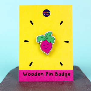 A wooden badge of a pink root vegetable with a smiling face and green leaf hair is shown on a yellow and pink backing board. At the bottom are the words Wooden Pin Badge.