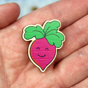 A wooden badge of a pink root vegetable with a smiling face and green leaf hair is shown held in an open hand.