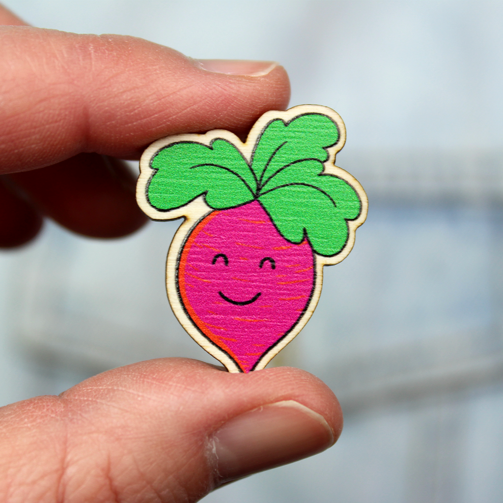 A wooden badge of a pink root vegetable with a smiling face and green leaf hair is held between finger and thumb in front of a denim jacket.