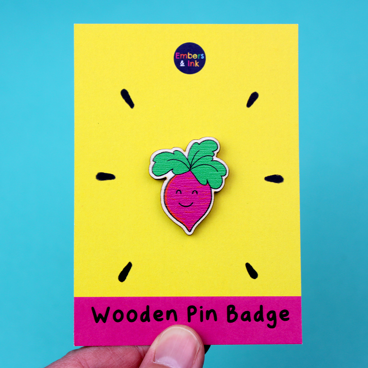 A wooden badge of a pink root vegetable with a smiling face and green leaf hair is shown on a yellow and pink backing board. At the bottom are the words Wooden Pin Badge.