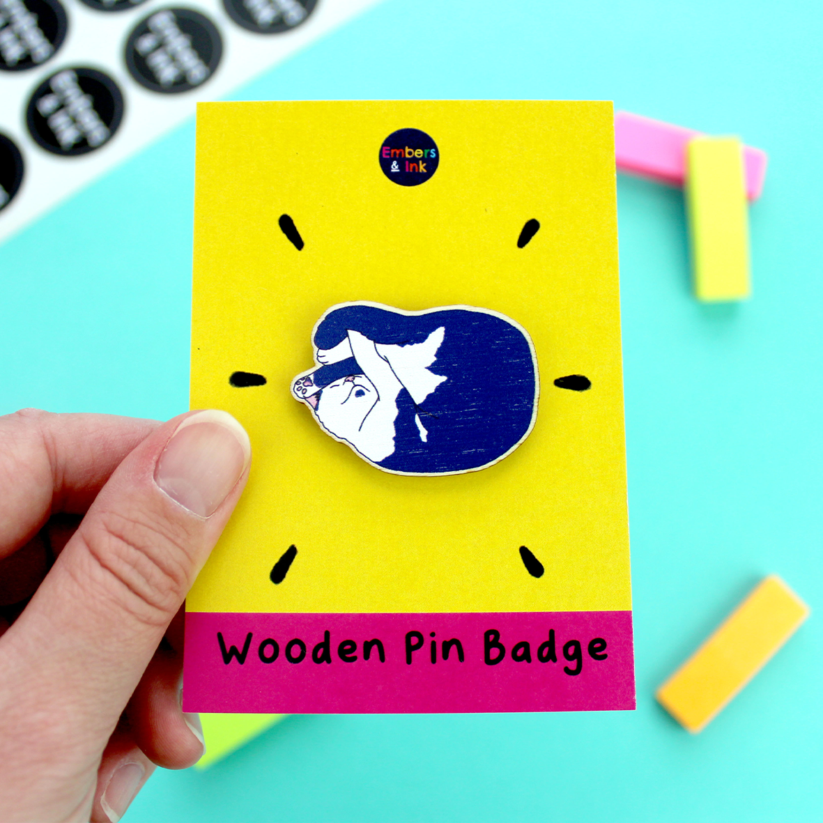 a hand holds a yellow and pink backing board that holds a wooden pin badge of a cat illustration. The cat is blue and white and laying in a cute way