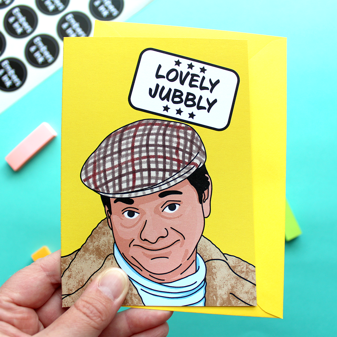 A hand holds a yellow card with an illustration of Del Boy from a well known 80s TV programme. Above him in a white box are the words ‘Lovely Jubbly’