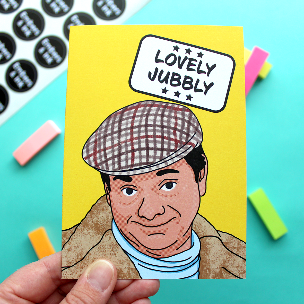 A hand holds a yellow card with an illustration of Del Boy from a well known 80s TV programme. Above him in a white box are the words ‘Lovely Jubbly’