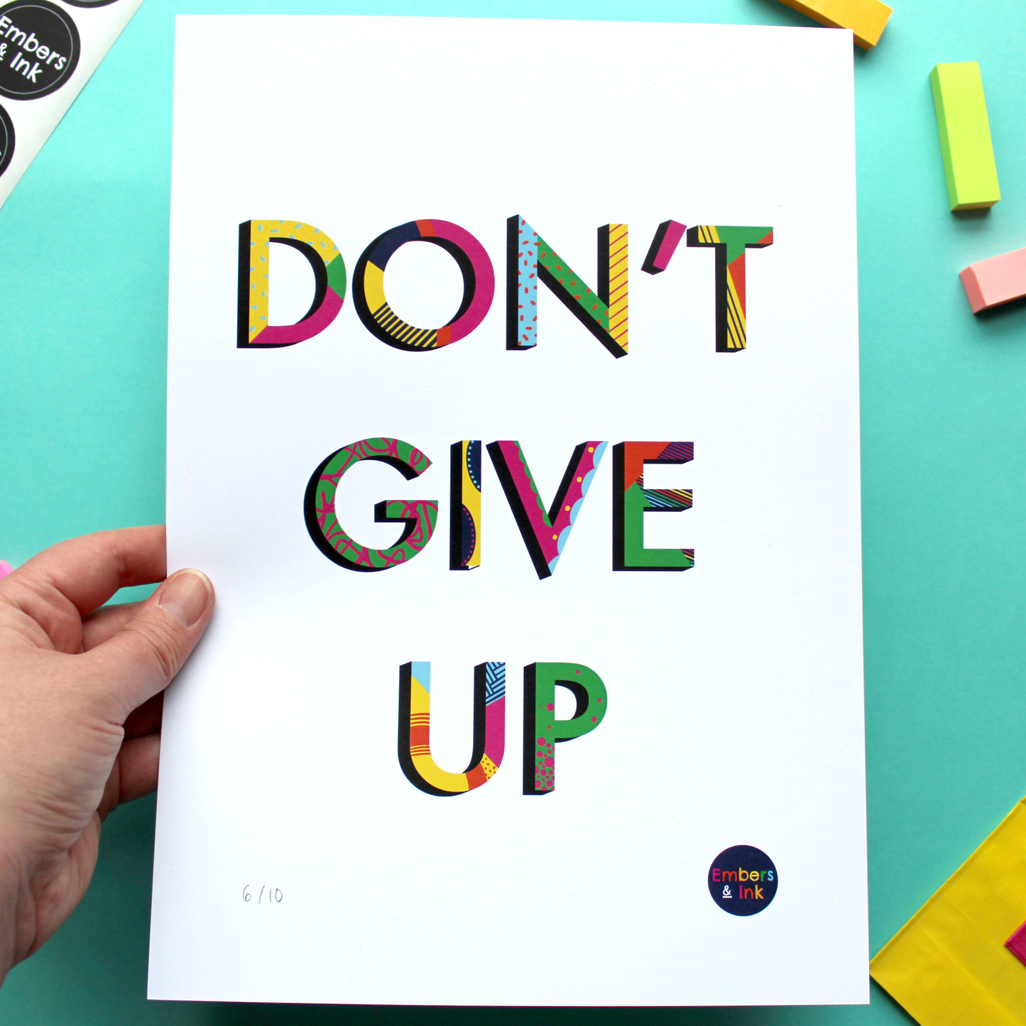 A hand holds an art print in portrait orientation features the coloured and patterned words Don't Give Up against a white background.