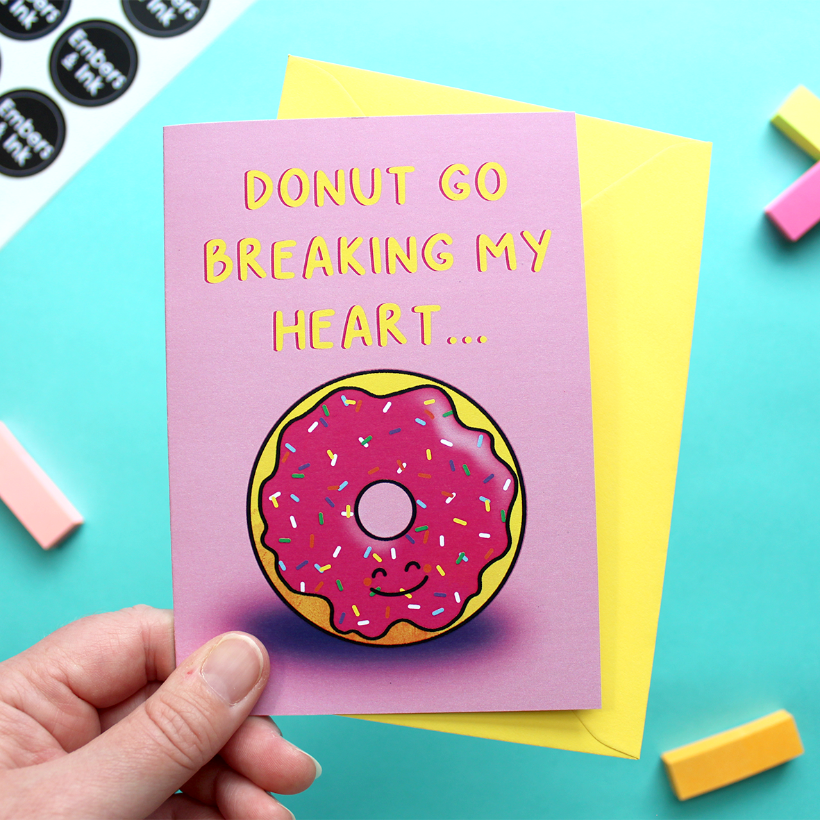 A hand holds a card that shows an illustration of a smiling iced donut, underneath the words 'donut go breaking my heart'. The background is pink.