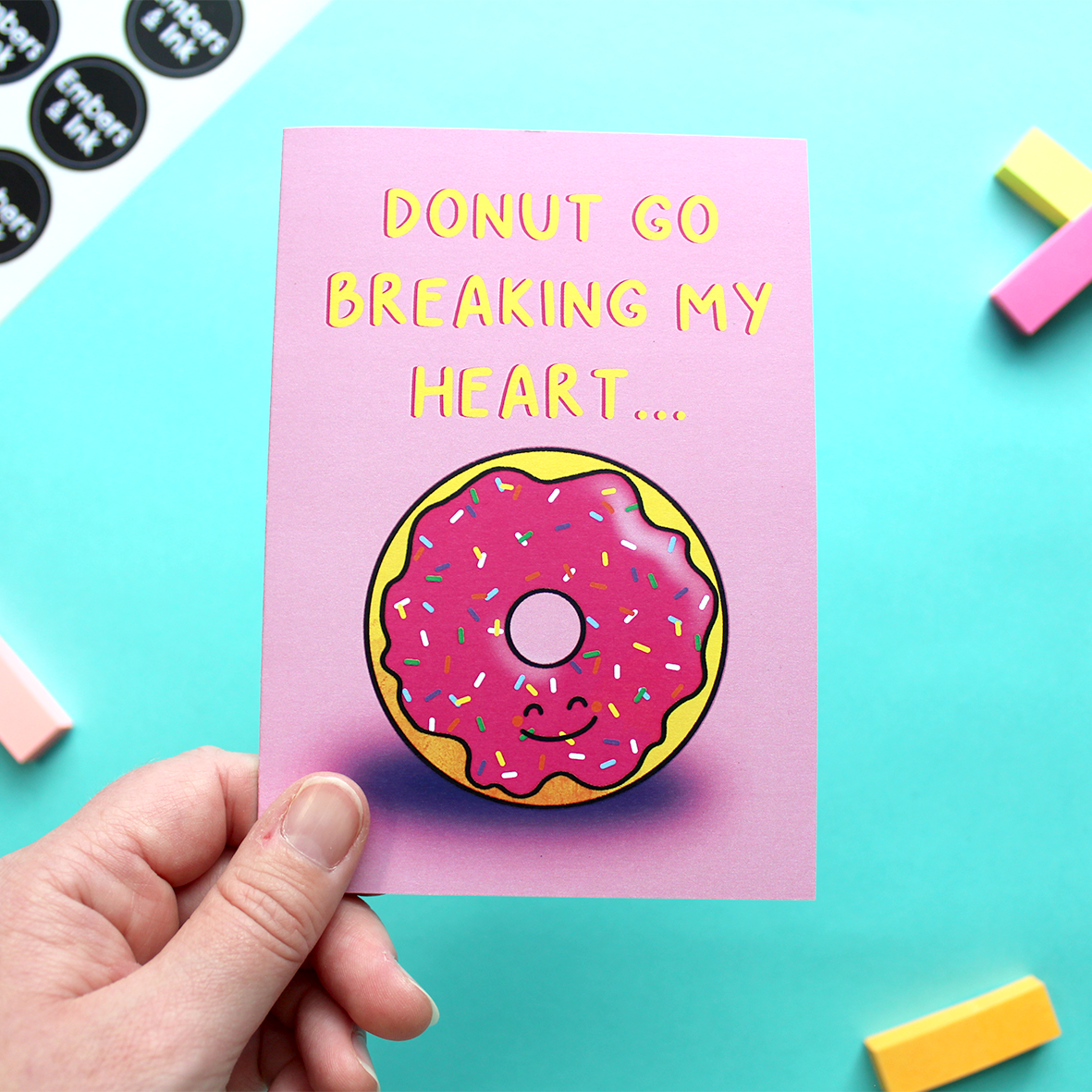 A hand holds a card that shows an illustration of a smiling iced donut, underneath the words 'donut go breaking my heart'. The background is pink.