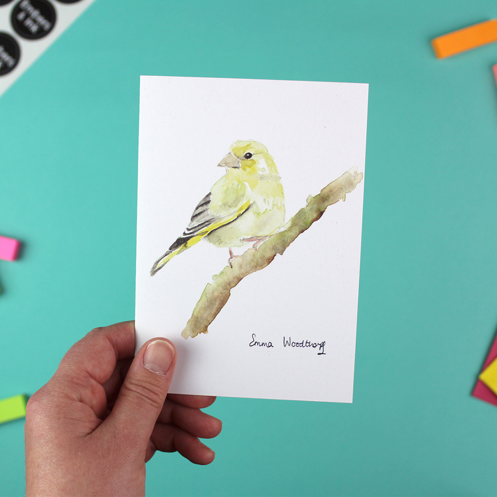 A hand holds a portrait-orientated image of a greenfinch bird in watercolour.