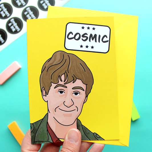 a hand holds a yellow card with an illustration of Rodney from a well known TV program underneath the word 'Cosmic' in a white box