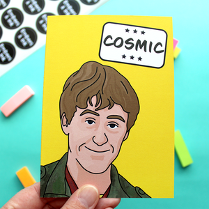 a hand holds a yellow card with an illustration of Rodney from a well known TV program underneath the word 'Cosmic' in a white box