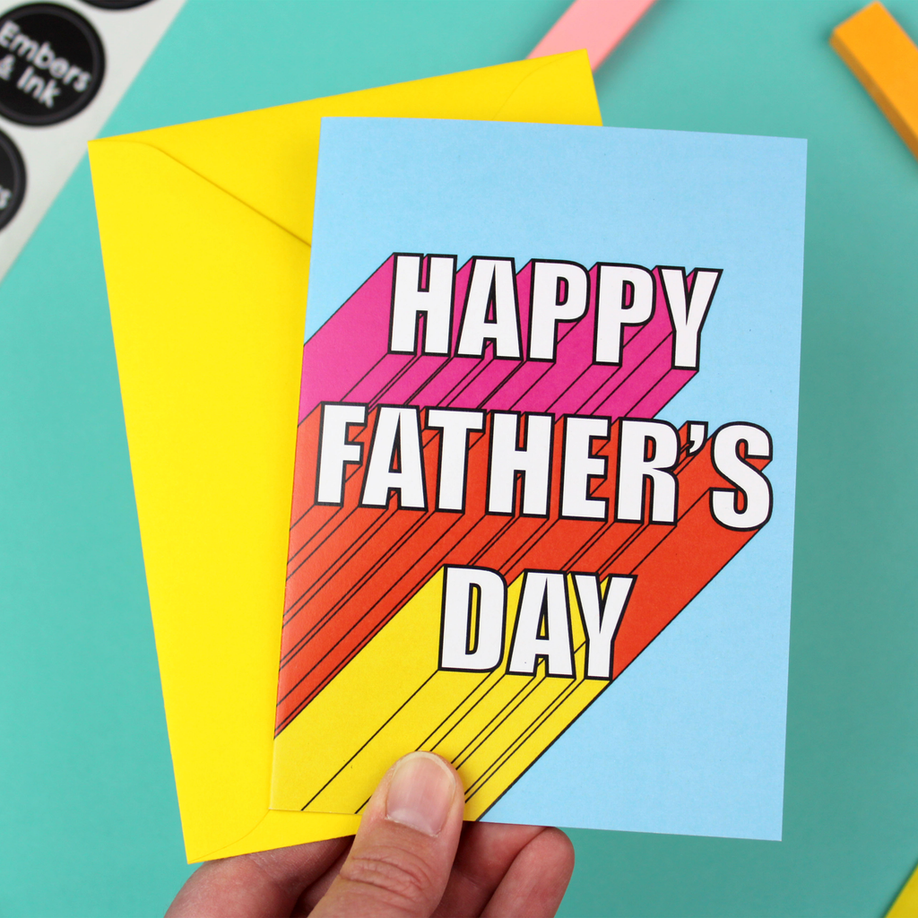 A hand holds a blue cards and a yellow envelope. The card has white writing that reads 'Happy Father's Day'. It has a 3D effect, with pink, orange and yellow colours.