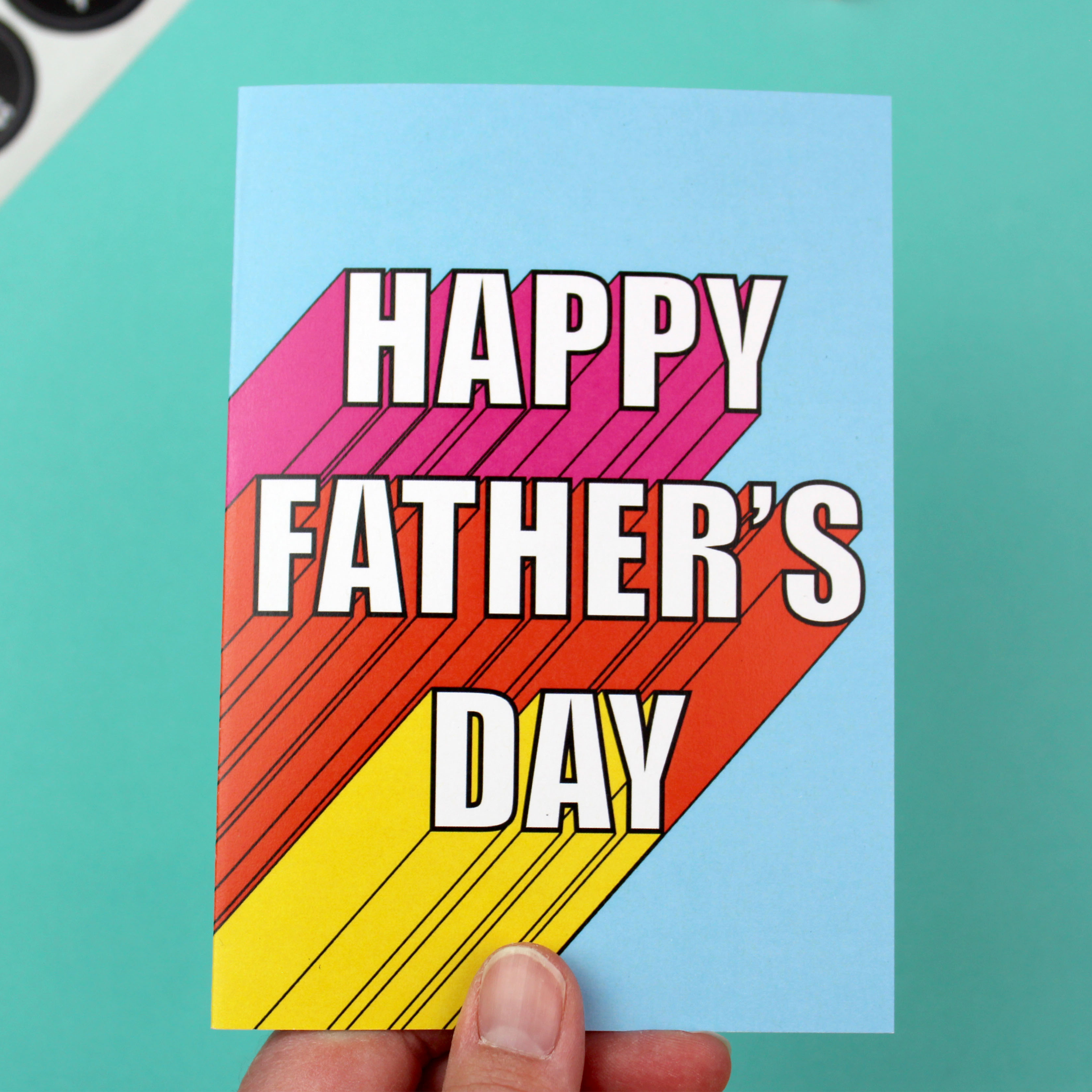 A hand holds a blue cards and a yellow envelope. The card has white writing that reads 'Happy Father's Day'. It has a 3D effect, with pink, orange and yellow colours.