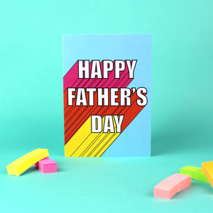 A blue card stands against a  green background. The card has white writing that reads 'Happy Father's Day'. It has a 3D effect, with pink, orange and yellow colours.