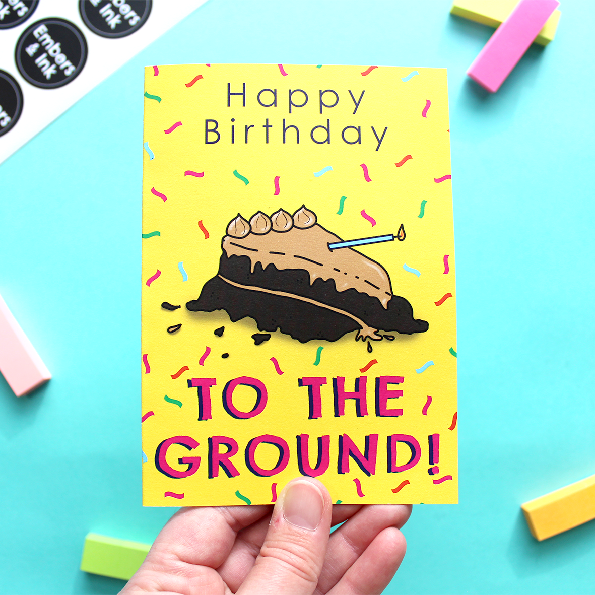 A hand holds the card which shows an illustration of a cake that's been thrown on the ground. We know this because the words on the card say 'Happy Birthday TO THE GROUND!'. It's taken from a song by The Lonely Island. The background of the card is yellow with multicoloured confetti 