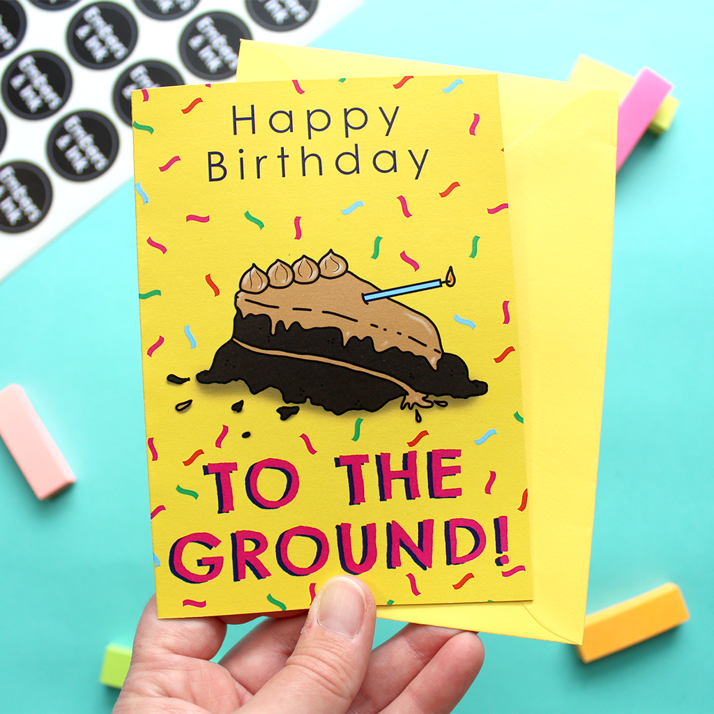 A hand holds the card which shows an illustration of a cake that's been thrown on the ground. We know this because the words on the card say 'Happy Birthday TO THE GROUND!'. It's taken from a song by The Lonely Island. The background of the card is yellow with multicoloured confetti 