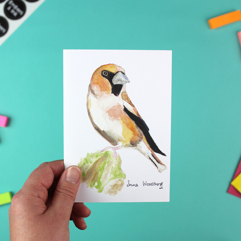 A hand holds a portrait-orientated A6 greetings card which features a watercolour image of the bird the Hawfinch.
