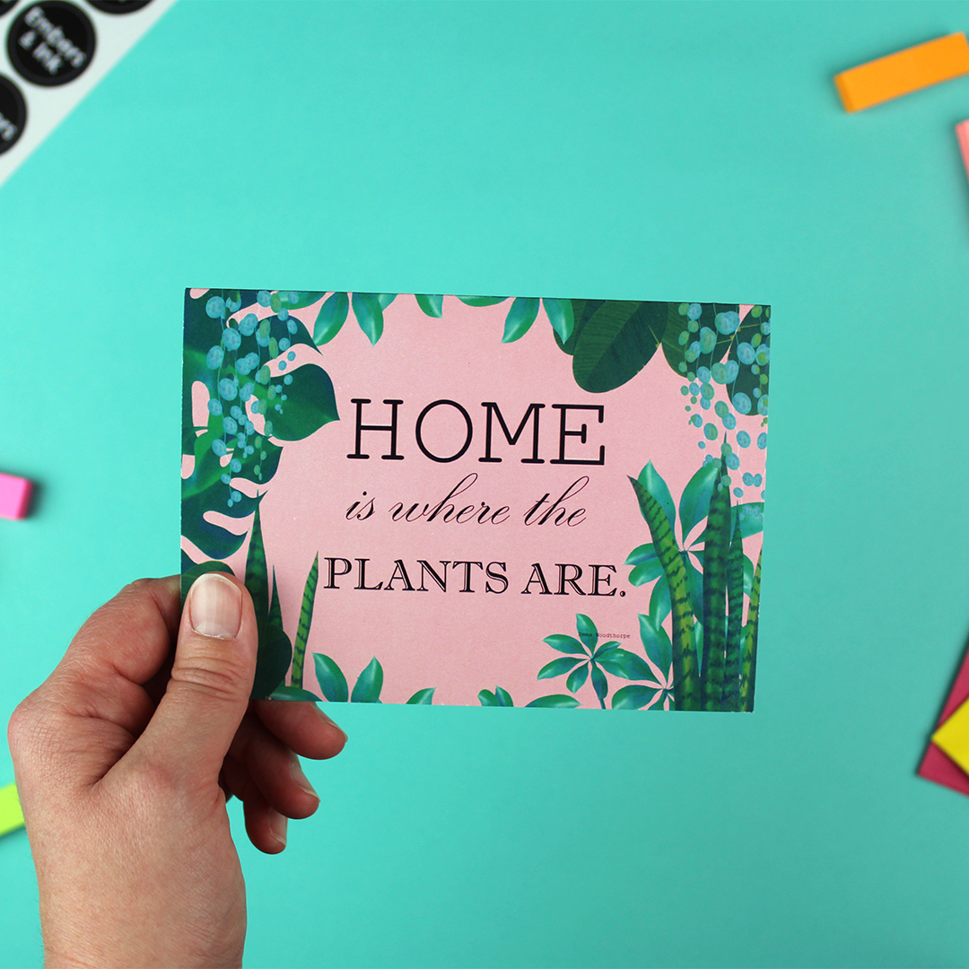 A hand holds a landscape orientated greetings card which has a border of house plants on a pink background. iside are the words 'home is where the plants are'