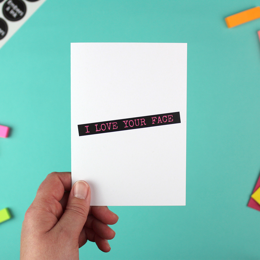 A hand holds a white card showing text in the style of a dymo-letter writer. The text readt 'I love your face'. The writing is pink with a black box background.