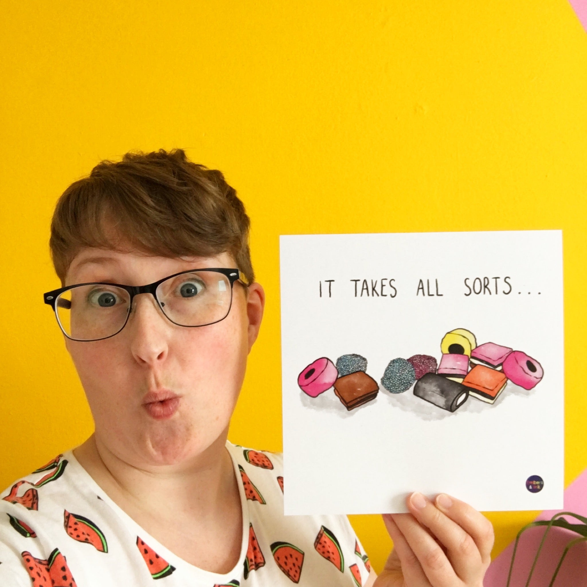A square art print with an illustration of a pile of All Sorts sweets is shown being held by Emma, owner of Embers and Ink. She is a 37 year old woman with short hair, and glasses, and standing in front of a yellow wall.. On the print, above the illustration, are the words 'it takes all sorts'