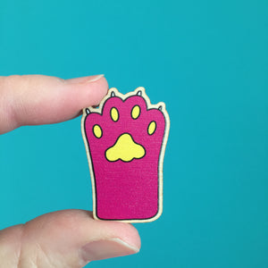 a pink paw wooden pin badge (with yellow pads and white claws) is held between a finger and thumb to show its size. it is approximately 37mm high and 25mm wide/