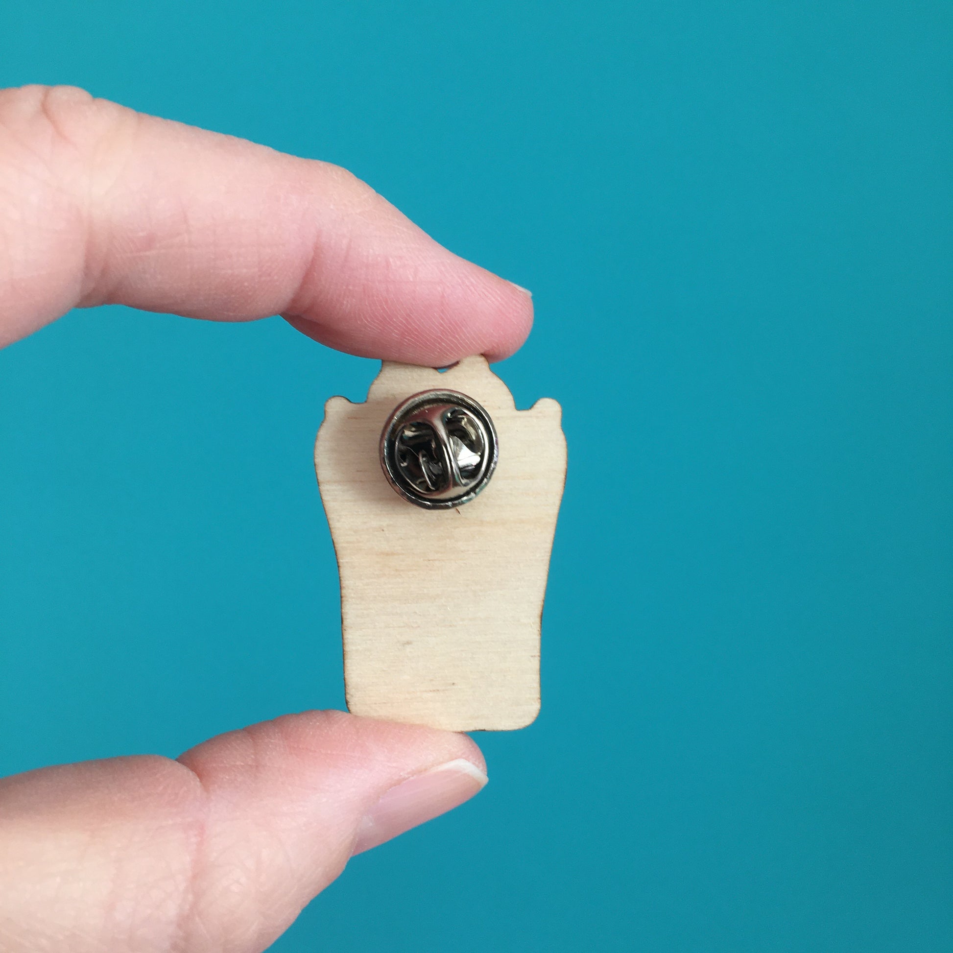 the back of the wooden pin badge is shown, to reveal the metal fastener with a metal butterfly clip.