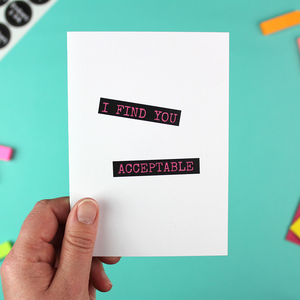 A hand olds a greetings card that says 'I find you acceptable' in pink text with a black box to imitate a dymo-gun effect. All on a white background.