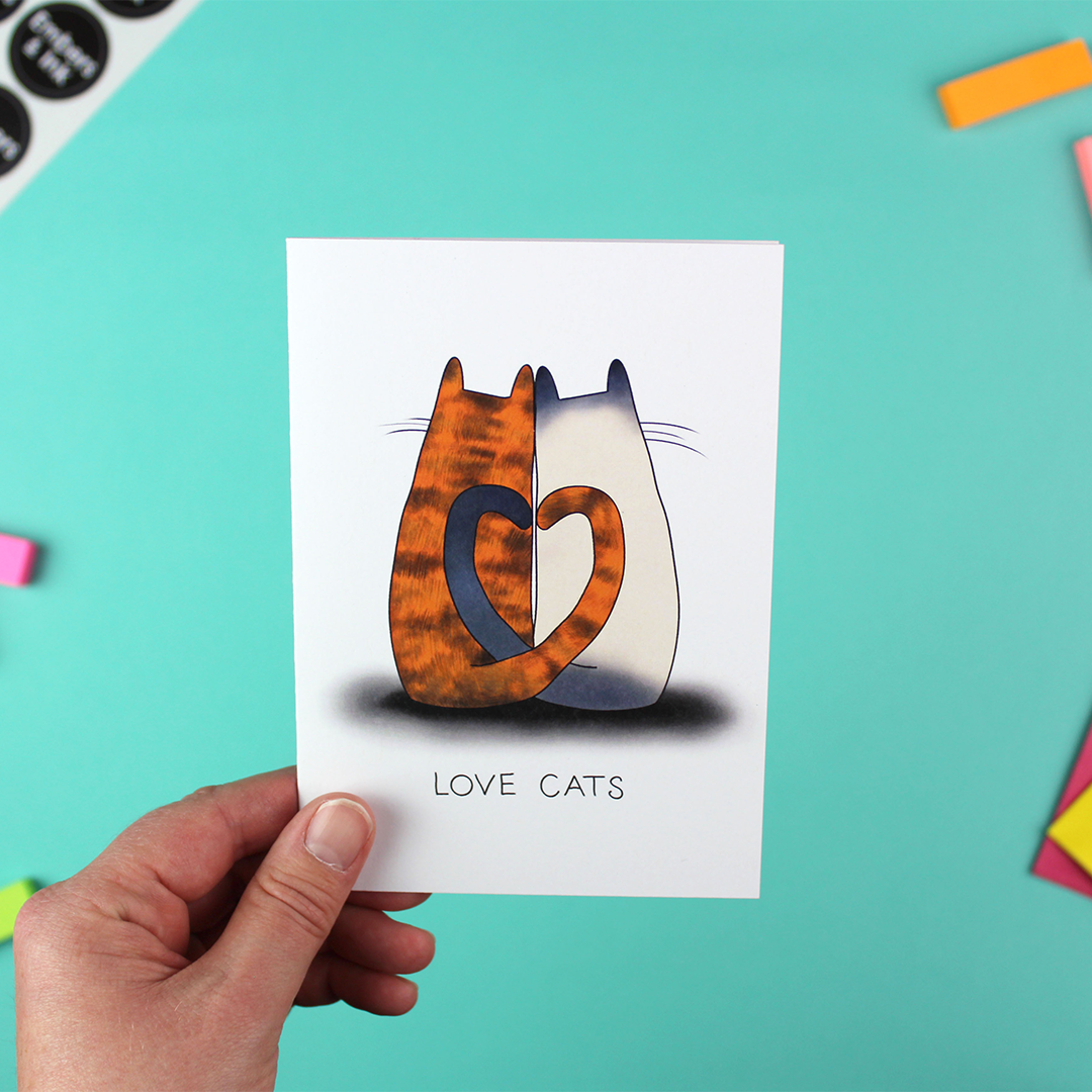 A hand holds a greetings card with an illustration of two cats leaning together with their tails entwined to form a heart. The left cat is a gingery colour and the right car is grey siamese-tye colouring with a beige body and grey tail and ears. Under the image are the words 'Love Cats'