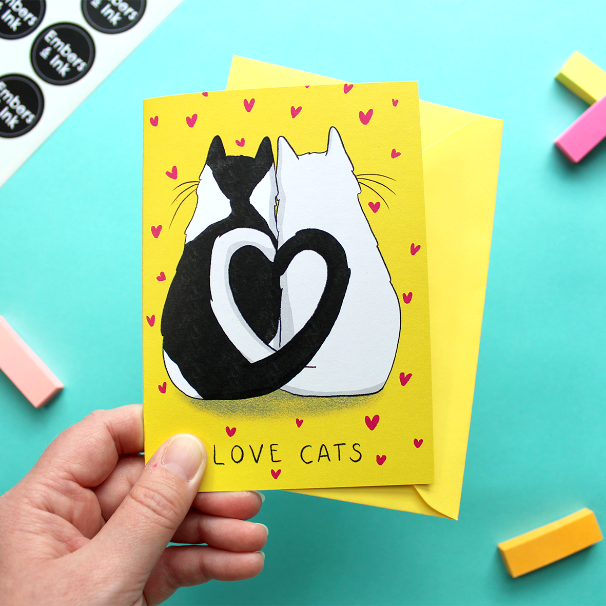 A hand holds a greetings card with an illustration of two cats facing away from us, with their tails entwined to make a heart shape. Below them are the words 'Love Cats'. The background of the card is yellow and the cats are surrounded by many small pink hearts.