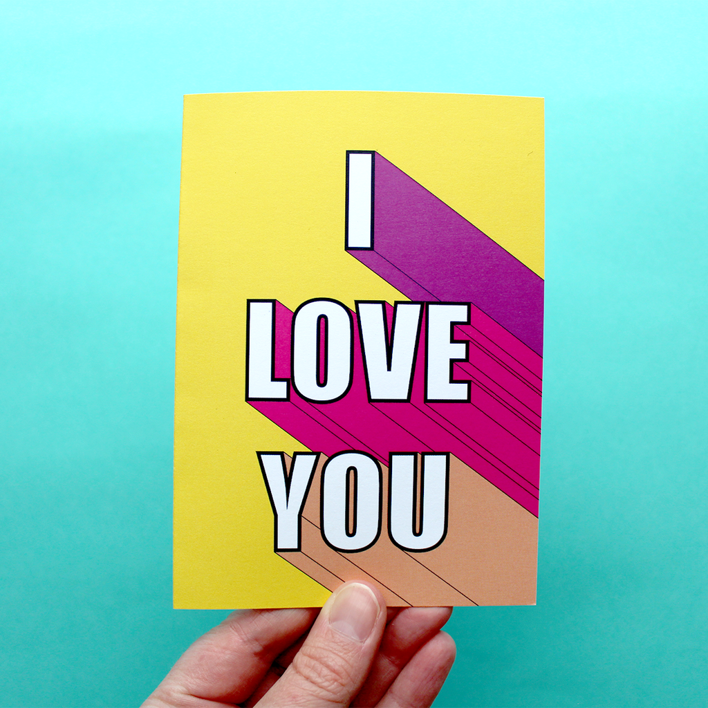 A hand holds an A6 greetings card in Portrait Orientation which has the 3D words 'I Love You' written in shades of pink on a yellow background.