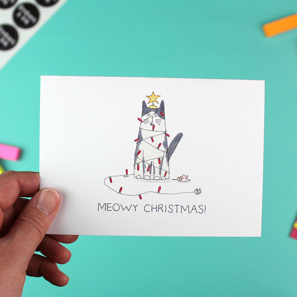 A hand holds a landscape orientated greetings card with an illustration of a black and white cat tangled in christmas lights with a star decoration on its head. Under it are the words 'Meowy Christmas'