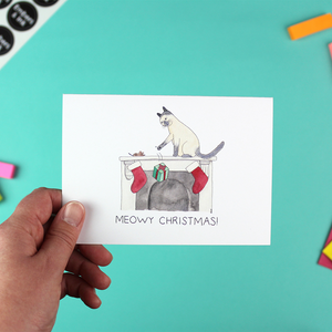 A hand holds a landscape orientated christmas card with an illustration of a cat knocking a present off a fireplace. Underneath are the words 'Meowy Christmas'