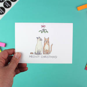 A hand holds a landscape orientated christmas card with an illustration of two cats sitting under the mistletoe. Underneath them are the words 'meowy christmas'