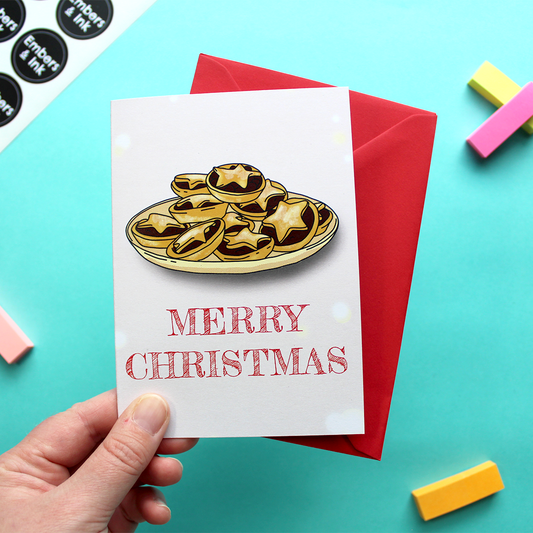 A hand holds a card that shows an illustration of a plate of mince pies above the words Merry Christmas. The text is in red and the background is pink.