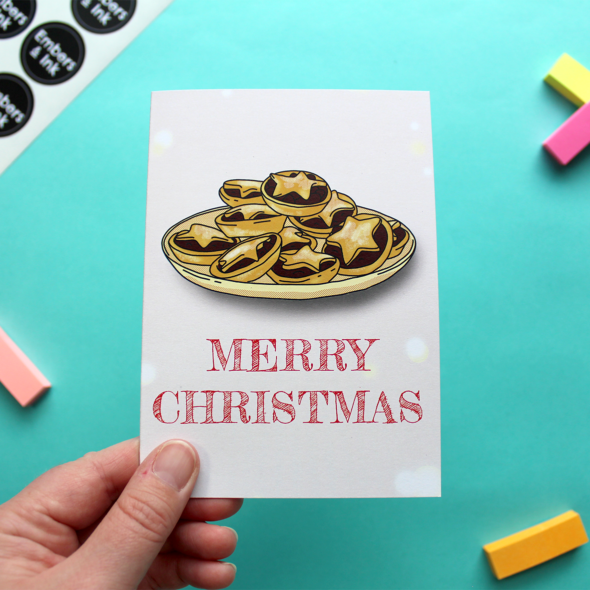 A hand holds a card that shows an illustration of a plate of mince pies above the words Merry Christmas. The text is in red and the background is pink.