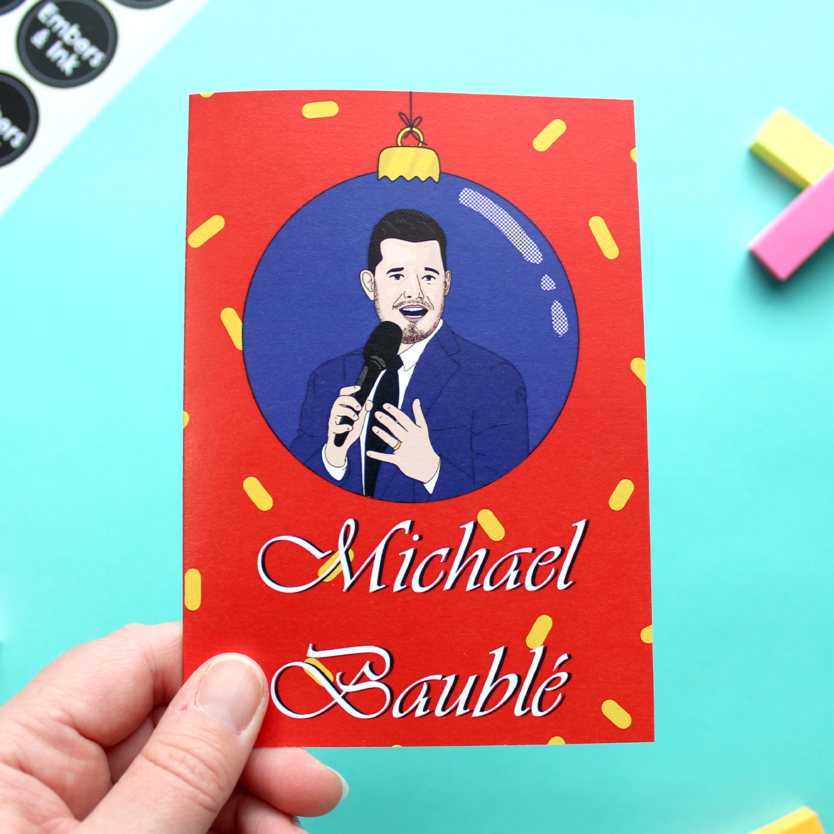 A hand holds a card that features an illustration of Michael Bublé within a blue bauble, above the words Michael Baublé. The background is red with yellow confetti-type shapes.