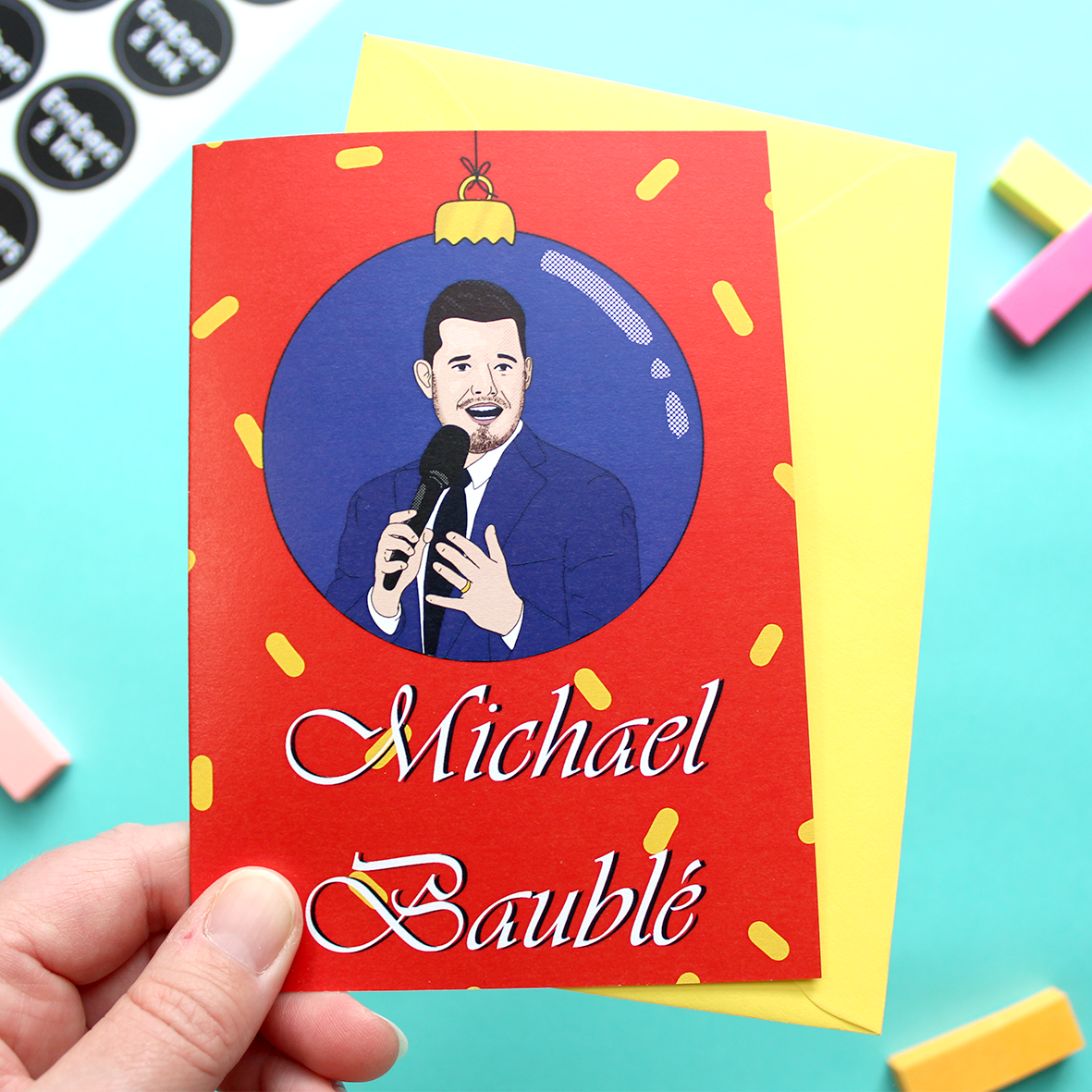 A hand holds a card that features an illustration of Michael Bublé within a blue bauble, above the words Michael Baublé. The background is red with yellow confetti-type shapes.