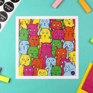 A square colourful poster is laying on a colourful desktop. It is full of cat illustrations with differing facial expressions. The cats are pink, yellow, green, blue and orange and they are like a bag full of jelly beans! There is a white border and in the bottom right corner is the Embers and Ink logo.