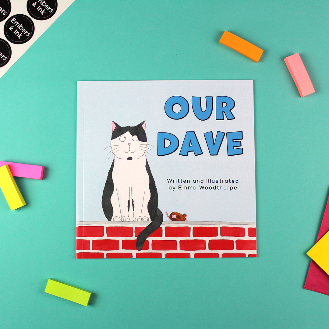A children's book called Our Dave by Emma Woodthorpe is laying on a colourful table. The front cover shows an illustration of a black and white cat sitting on a wall next to a toy mouse.