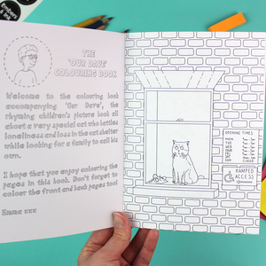 A hand holds open the colouring book to show the inside cover - which has a welcome note-  and the first page which shows a cat looking out of a window. both pages are black and white and ready to be coloured in.