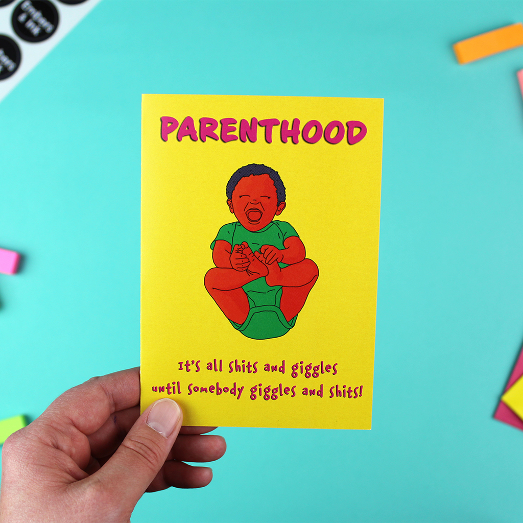 A hand holds a bright yellow card with an illustrated baby in the centre, with orange skin, blue hair and a green romper suit. aroung it are the words: 'parenthood: it's all shits and giggles until somebody giggles and shits'