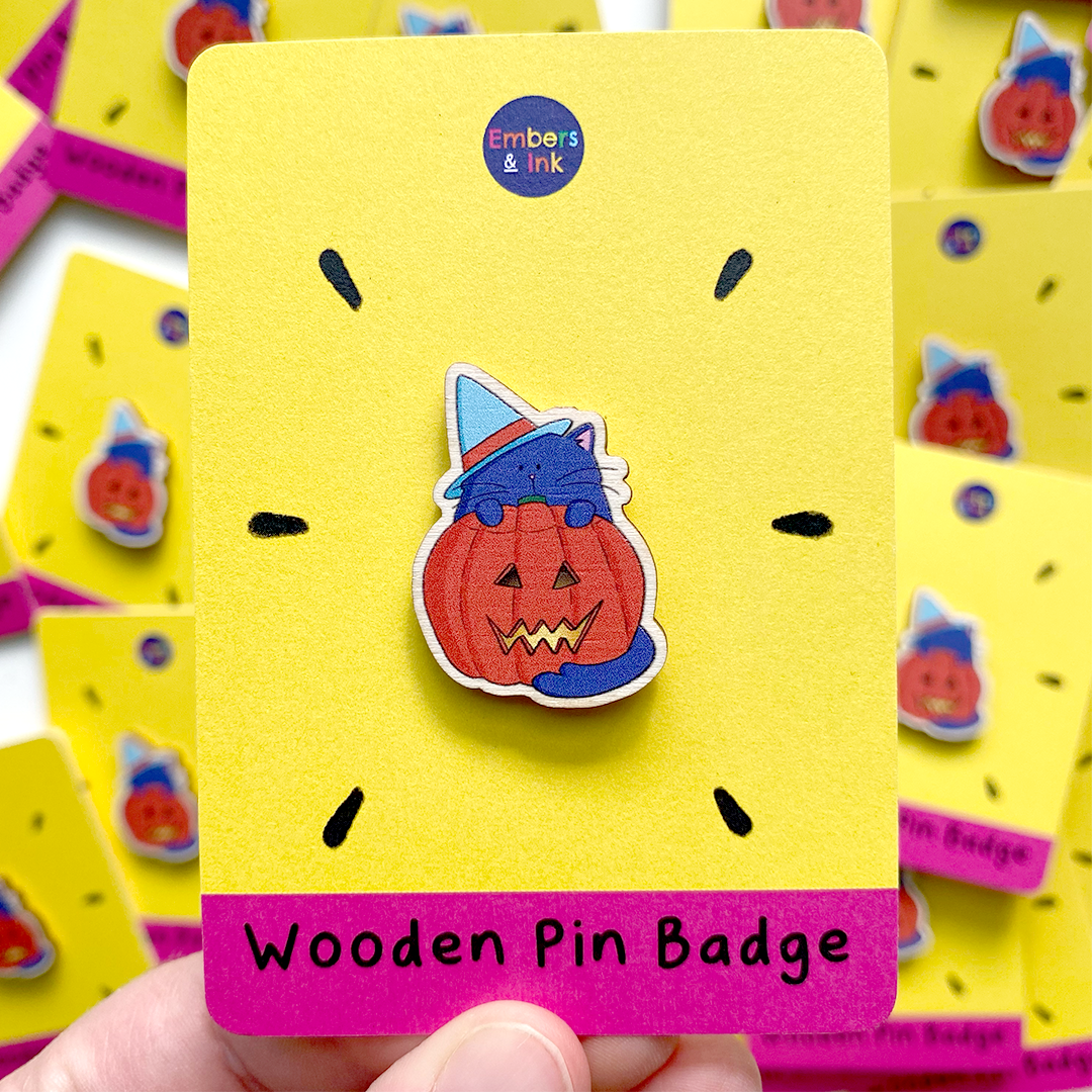 a wooden pin badge featuring an illustrated purple cat wearing a blue witches hat and standing behind a lit spooky halloween pumpkin is shown on a yellow and pink cardboard display backer that says 'Wooden Pin Badge' at the bottom/